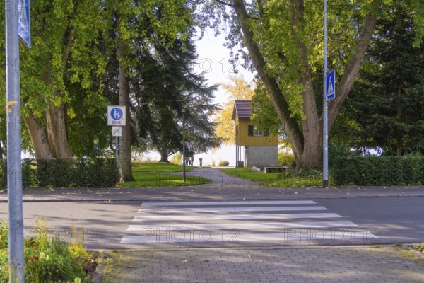 A pedestrian crossing leads to a park with tall trees and an autumn backdrop, Überlingen, Lake Constance, Germany