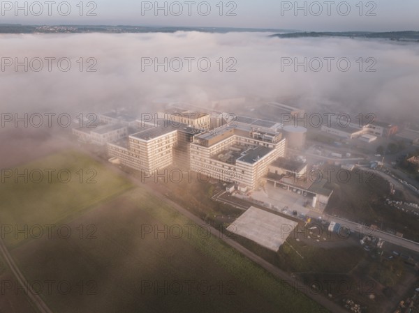 Large industrial buildings surrounded by fields under a thick layer of fog, new hospital health campus building, Calw, Germany