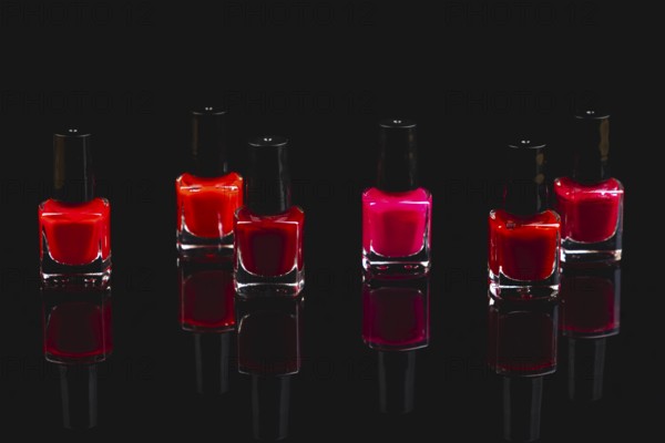 Six bottles of nail polish in shades of red and pink on a black surface with reflection
