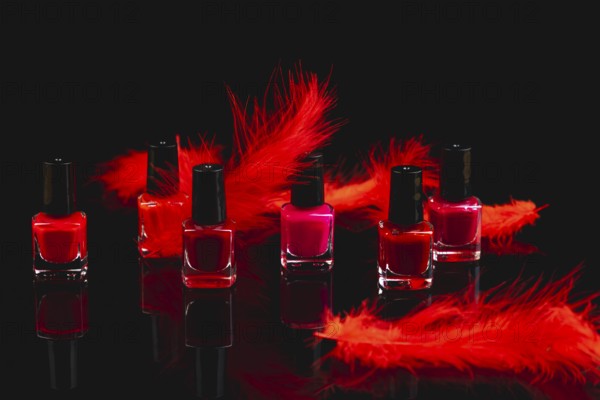 Nail polish bottles in shades of red and pink with red feathers on a black surface with reflection