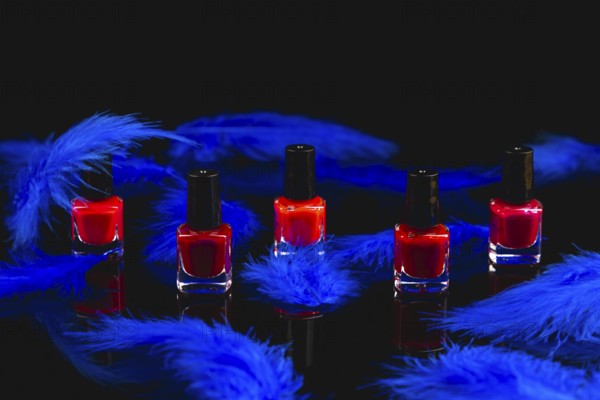 Red nail polish bottles surrounded by blue feathers on a black surface with reflection