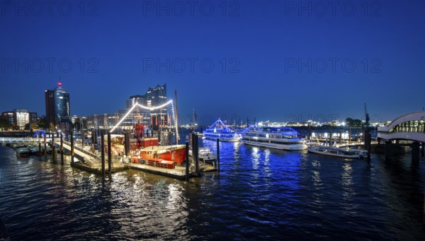 View of illuminated ships on the Elbe promenade with restaurant Das Feuerschiff LV 13, City Sporthafen, behind Speicherstadt with Elbphilharmonie and Columbus Haus, blue hour, Hamburg, Germany