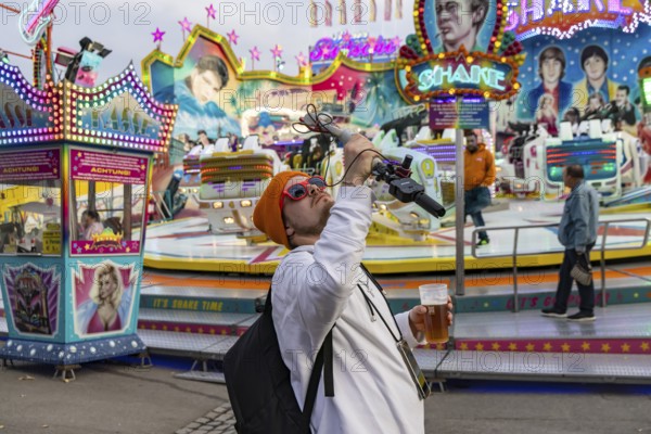 Influencer with live camera and beer in front of a fairground ride. Cannstatter Volksfest at the Wasen. Bad Cannstatt, Stuttgart, Baden-Württemberg, Germany