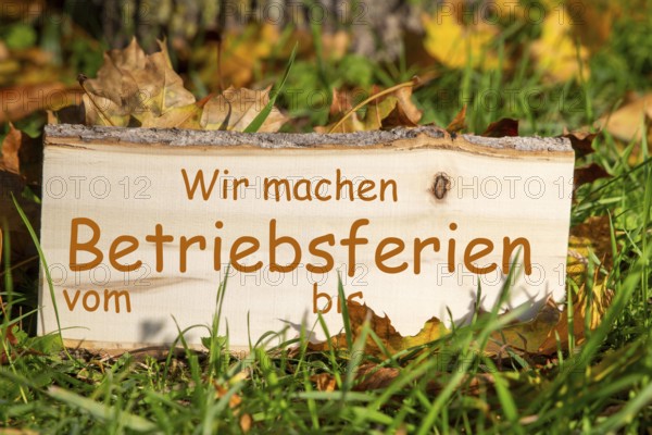Company holidays autumn: Wooden sign with added text WIR MACHEN BETRIEBSFERIEN VOM/BIS in a meadow with autumn leaves