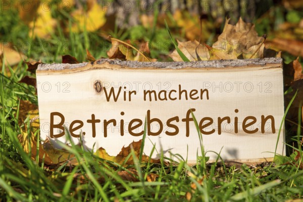Company holidays autumn: Wooden sign with added text WIR MACHEN BETRIEBSFERIEN in a meadow with autumn leaves