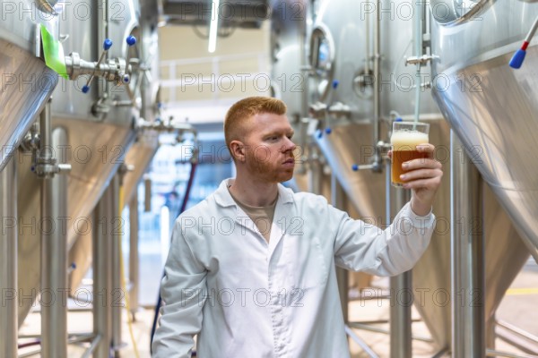 Brewmaster in lab coat inspects a glass of golden craft beer in a modern brewery, assessing color, clarity and foam as stainless steel tanks and equipment operate in the background