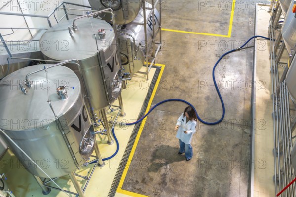 Woman scientist in a white lab coat monitoring stainless steel fermentation tanks in a modern brewery, ensuring quality control during the beer production process