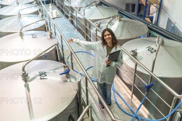 Woman wearing a white lab coat and holding a clipboard, standing on a metal gangway, showing a row of large stainless steel fermentation tanks in a modern brewery