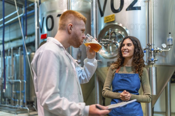 Brewery team performs quality control tasting and discussion beside stainless steel fermenters, brewmaster and technician inspecting beer and recording production notes