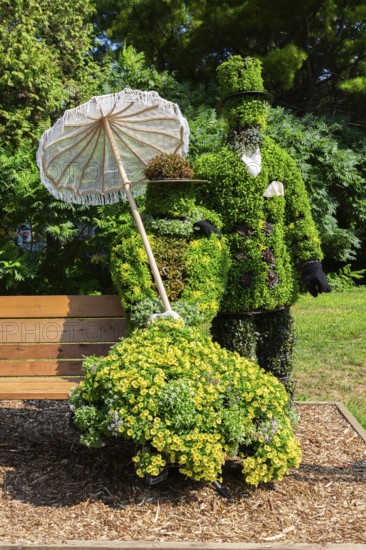 Live plant sculpture of Victorian man standing and woman with sun shade umbrella sitting on wooden park bench in summer, Ile des Moulins, Old Terrebonne, Quebec, Canada
