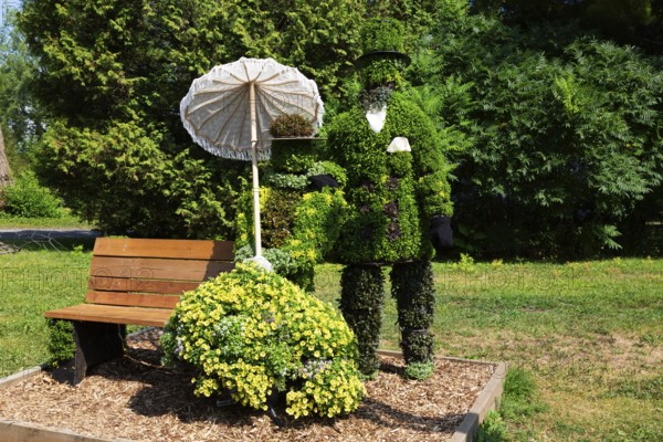 Live plant sculpture of Victorian man standing and woman with sun shade umbrella sitting on wooden park bench in summer, Ile des Moulins, Old Terrebonne, Quebec, Canada