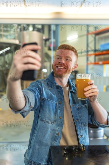 Smiling red haired man in a denim jacket takes a selfie with his smartphone while holding a glass of craft beer in a modern brewery among stainless tanks and equipment