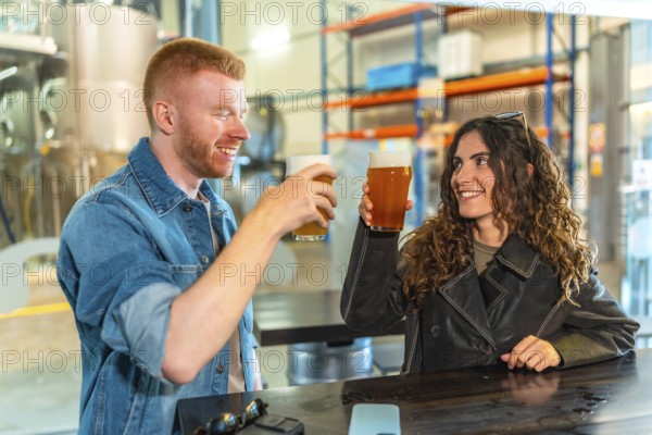 Young man and woman clinking craft beer glasses and smiling at a modern microbrewery bar, enjoying a casual date and social celebration amid industrial fermenters and warm lighting