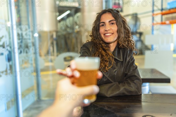 Woman smiling and raising a glass of frothy craft beer in a modern brewery, toasting an unseen companion across a wooden table during a relaxed, social weekend outing