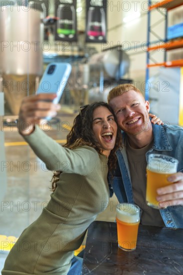 Happy young couple posing for a selfie with a smartphone, holding glasses of craft beer, and celebrating together inside a vibrant beer factory or microbrewery, having fun