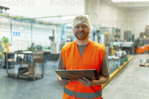 Male worker with a beard, wearing a reflective safety vest and hairnet, is confidently smiling at the camera while holding a clipboard in a bustling food and beverage production facility