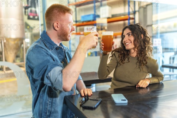 Happy young couple toasting craft beers and laughing together inside a modern brewery tasting room, enjoying a relaxed date and quality time amid industrial chic surroundings