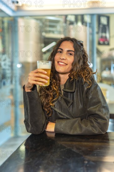 Smiling woman with curly hair wearing a leather jacket, happily holding a glass of pale beer while relaxing at a brewery bar, industrial tanks visible in the background