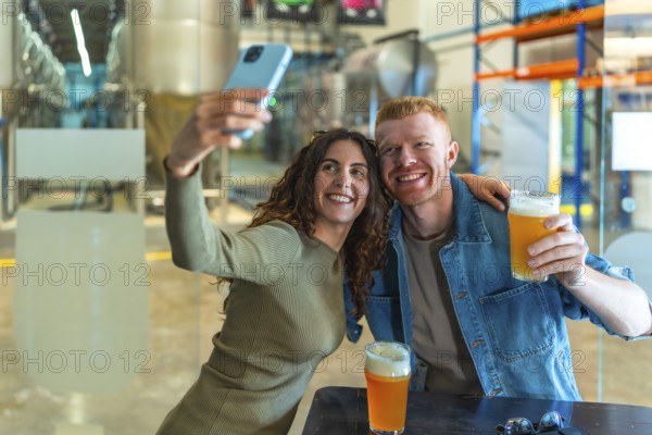 Young couple smiling for a selfie with craft beers in hand at a modern brewery tasting room, enjoying a casual weekend hangout, tasting and sharing the experience