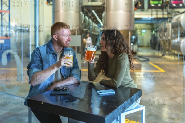 Couple sitting at a table in a modern craft beer factory, drinking and communicating while enjoying a brewery tour experience with large fermentation tanks in the background
