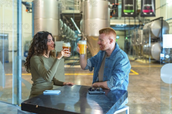 Young couple enjoying craft beer at a table, making a toast and smiling inside a modern beer factory with large stainless steel tanks in the background