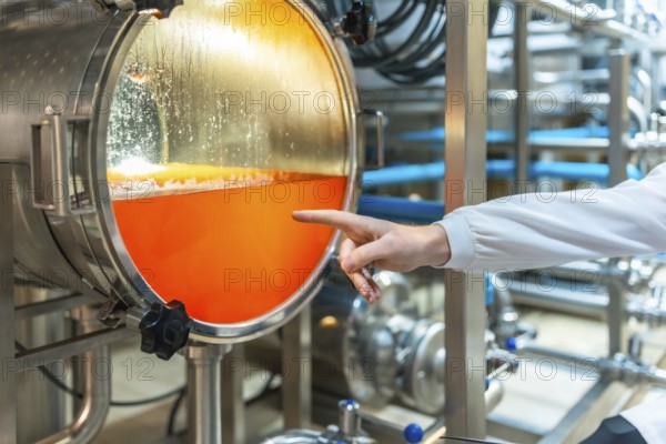 Brewery worker checking the vibrant orange liquid inside a stainless steel fermentation tank with a clear viewing window, observing the brewing process in a modern industrial beer factory
