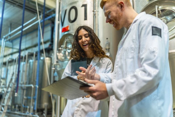 Brewery colleagues smiling and discussing production notes on a clipboard, standing in a beer factory with large stainless steel fermentation tanks in the background