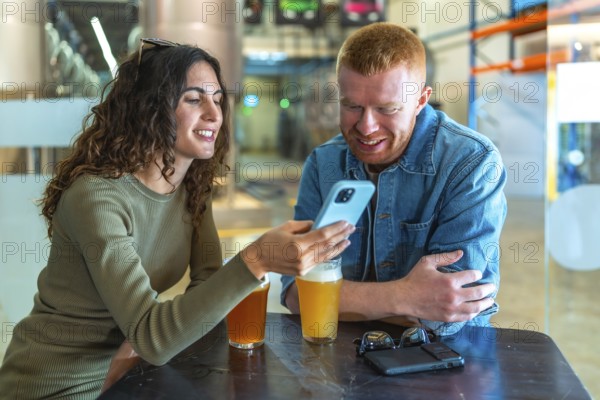 Young couple sitting at a table in a brewery or beer factory, having a conversation and sharing content on a smartphone while enjoying refreshing glasses of craft beer