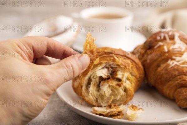 Croissant on white plate with hand on brown concrete background and linen textile, cup of coffee, side view, close up, selective focus