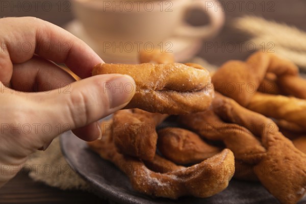 Crunchy biscuit Brushwood cookies with hand sprinkled with powdered sugar on brown wooden background and linen textile, cup of coffee, side view, close up, selective focus