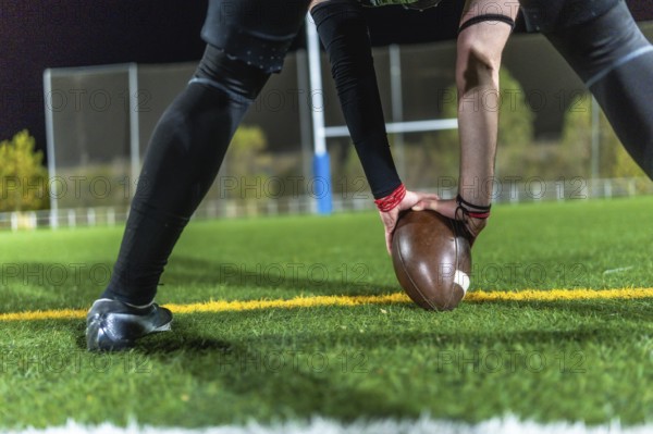 American football player in dark uniform with arm sleeves placing a brown ball on green turf at night, low angle close up capturing focus before kickoff