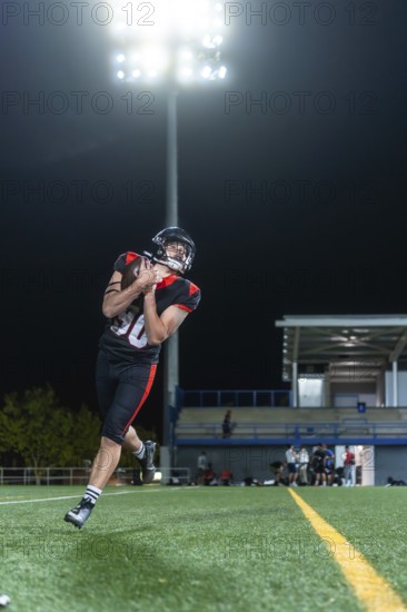 American football player in uniform and helmet catching the football on a green turf field at night under bright stadium lights, with blurred spectators in the stands
