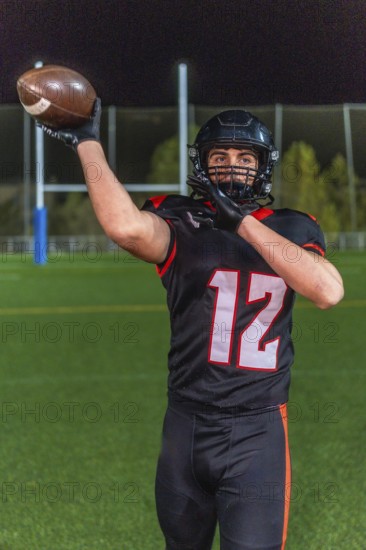 American football player in helmet and number 12 uniform holds the ball up on a green grass field under night stadium lights, poised and ready to pass or play