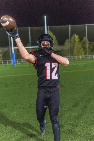 American football player wearing a black uniform with number 12 and helmet, extending arm to catch a brown football on a green field under stadium lights during an evening game