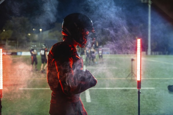 American football player standing on a dark field at night in helmet and pads, framed by red and blue smoke and dramatic lighting, projecting energy, determination and readiness