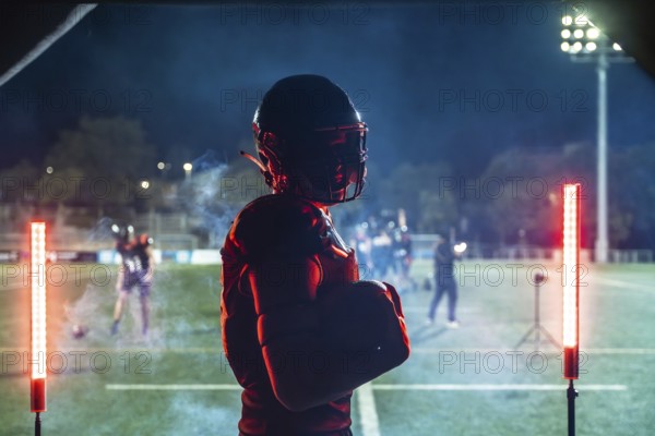 American football player in full gear holding a ball, standing confidently on a floodlit stadium field at night, intensely focusing on the upcoming game or practice