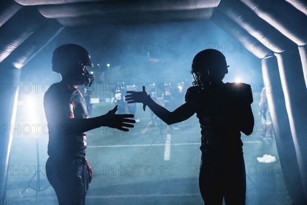 American football players in silhouettes high fiving each other inside a smoky stadium tunnel at night, getting ready for a game as teammates wait on the field