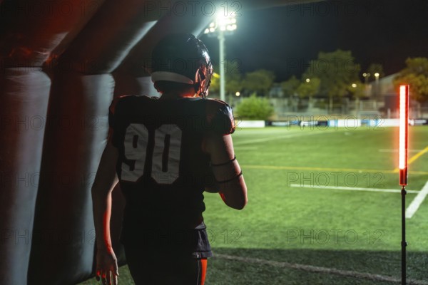 American football player in jersey number 90 emerges from an inflatable tunnel onto a floodlit stadium field at night, low angle back view capturing power and anticipation