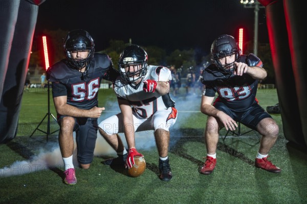 Three determined american football players, one holding the ball, emerging from an inflatable tunnel onto a stadium field at night, ready for game action