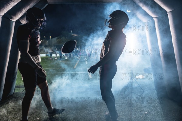 Two american football players wearing helmets and uniforms emerge from a tunnel onto stadium field at night, a football hovering between them in a misty atmosphere, ready for a game