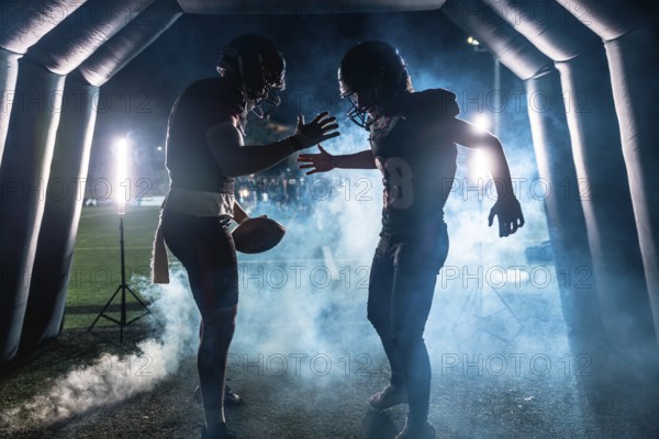 American football players, one holding a ball, emerging from a smoky stadium tunnel at night, preparing for a game and high fiving in a moment of teamwork and readiness