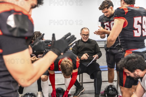 American football coach smiling, discussing strategy, and preparing for a game with diverse male players dressed in uniforms and protective gear inside a locker room