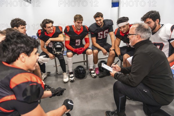 American football coach kneeling, explaining game strategy to a team of young athletes in full uniform, sitting on a bench in a locker room, focusing on teamwork and upcoming plays