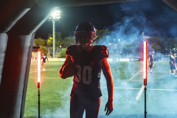 American football player in helmet and uniform bursts from a smoke filled tunnel onto a floodlit field at night, clutching the ball, poised and ready for game action and competition