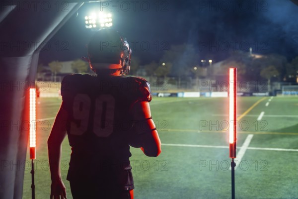 American football player in full uniform and helmet emerging from a tunnel onto a floodlit stadium field at night, holding the ball and poised to charge into play