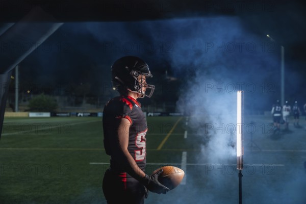 American football player is holding a ball, wearing a helmet and uniform, and standing on a playing field at night with smoke effect and stadium lights