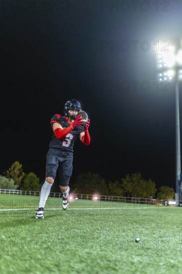 Male football player making a dramatic evening catch under stadium lights on green turf, showcasing focus, athleticism and dynamic movement during a competitive night game