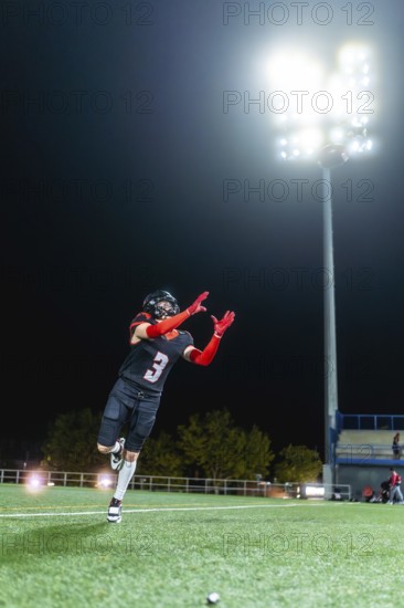 American football player in uniform with a number 3 catching a ball on a lit artificial turf field at night, demonstrating athleticism and focus during practice