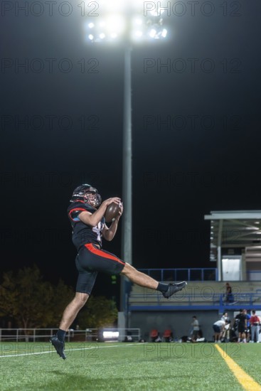 American football player in uniform and helmet leaping to catch the ball mid air on a floodlit stadium field at night, demonstrating athleticism and skill
