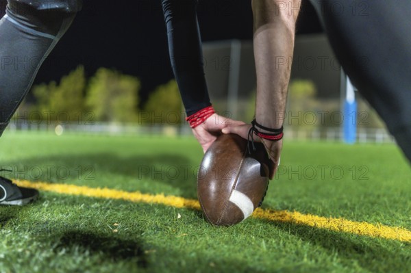 Football player positioning brown leather ball on yellow line on green grass, hands steadying ball before kickoff during evening match, focus and preparation on the field